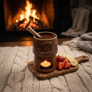 May include: A brown ceramic fondue pot with a light-coloured wooden stirring stick, featuring the text "FONDUE MOMENTS" and a small heart icon. The pot sits on a wooden board with sliced bananas and strawberries, illuminated by a tea light candle.