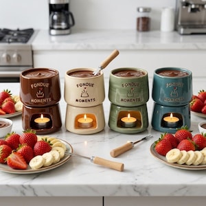May include: Four ceramic fondue pots in brown, cream, sage green, and teal, each featuring the text FONDUE MOMENTS and a small heart icon. Each pot sits on a base with a lit tea light candle inside. Two metal forks with wooden handles are included.