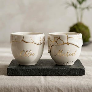 May include: Two off-white ceramic cups featuring gold-coloured crack patterns along the upper rims. The left cup is inscribed with the name Chloe in gold script, and the right cup features the name Micheal in gold script, both in a cursive font.