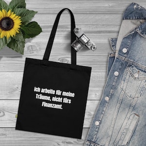 Puede incluir: Bolso tote negro con texto blanco "Ich arbeite für meine Träume, nicht fürs Finanzamt." Una cámara vintage cuelga del asa del bolso. Un girasol y una chaqueta vaquera están en el fondo.