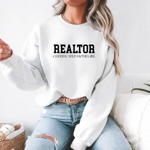 May include: White sweatshirt with the word "REALTOR" in bold, black capital letters, and the text "CERTIFICATED HOMEGIRL" below it. The sweatshirt is paired with blue jeans.