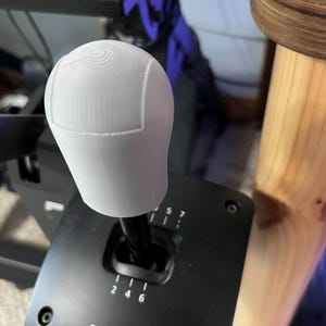 May include: A white, 3D-printed gear shift knob with a textured surface, mounted on a black Fanatec shifter. The knob has a rounded top with a circular design. The shifter shows gear positions.