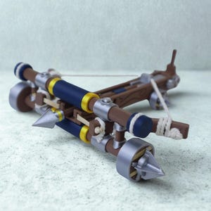 May include: A miniature crossbow with a brown wooden frame, detailed with blue and gold accents. The crossbow has a silver arrowhead and a white string. The design includes silver metal accents and a detailed, realistic appearance.