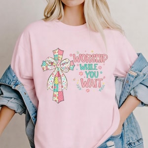 以下が含まれることがあります： 明るいピンク色のスウェットシャツ。カラフルな十字架のデザインと「Worship While You Wait」の文字が特徴です。十字架はパステル模様とリボンで装飾されています。デザインには花のアクセントが含まれています。