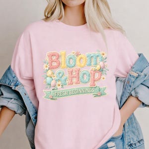 Könnte beinhalten: Ein hellrosa Sweatshirt mit dem Schriftzug "Bloom & Hop" in bunten, strukturierten Buchstaben. Das Design umfasst Ostereier und Blumen, mit dem Satz "Fresh Beginnings" auf einem Banner darunter. Eine Jeansjacke ist über den Schultern getragen.