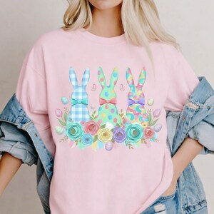 May include: Pale pink sweatshirt with a design of three Easter bunnies, each with a bow tie and surrounded by flowers. The bunnies have different patterns: blue gingham, green polka dots, and a colourful confetti design. Pastel eggs and flowers complete the spring theme.
