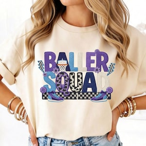 May include: Cream-colored t-shirt with the words "BALLER SQUAD" in purple, blue, and black. The design includes basketballs, a basketball hoop, flowers, and sneakers. The letters have a 3D effect.