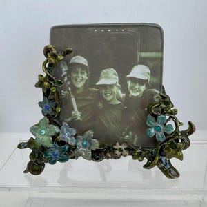 May include: A decorative photo frame with a floral design. The frame features a black and white photo and is adorned with green, blue, and gold-colored flowers and leaves. The frame is made of metal and has a glossy finish.