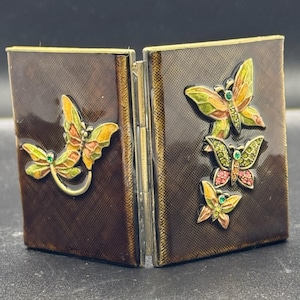 May include: A hinged, brown metal case with a textured surface. The case is decorated with colorful butterfly embellishments in shades of yellow, green, and pink, each with a small gemstone. The case is open, revealing the interior.