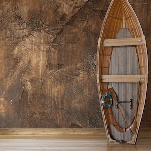 May include: A wooden rowing boat with a lifebuoy and oars inside, standing upright against a brown textured wall in a room with a wooden floor.