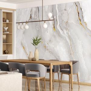 Puede incluir: Un comedor moderno con un papel tapiz de mármol blanco con vetas doradas. Una mesa de comedor de madera con seis sillas se coloca frente al papel tapiz. La mesa está hecha de madera clara y tiene un diseño simple. Las sillas están tapizadas en tela gris y tienen un aspecto elegante y moderno.
