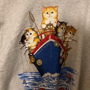 Puede incluir: Una sudadera gris claro con un estampado gráfico de un barco pesquero azul y rojo sobre el agua. El barco está ocupado por cinco gatitos, uno al timón, otro con una red de pesca y otros con cañas de pescar. El texto dice Catch of the Day.
