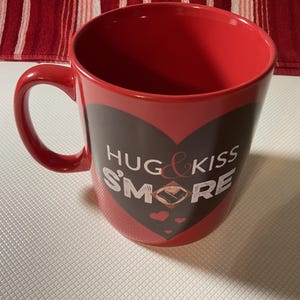 May include: A red ceramic mug with a black heart design. The mug has the words "HUG & KISS S'MORE" printed on it. The mug has a red handle and a red interior.