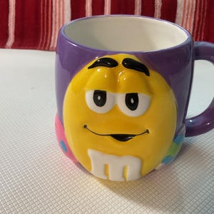 May include: A ceramic coffee mug with a purple exterior and a white interior. The mug features a large, smiling yellow M&M'S character on the front. The handle and the rim of the mug are purple. The mug is on a white surface.