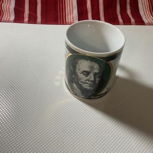 May include: A white ceramic mug featuring a portrait of a man. The portrait is framed with a gold border and set against a green background. The mug has a black band at the top and bottom.