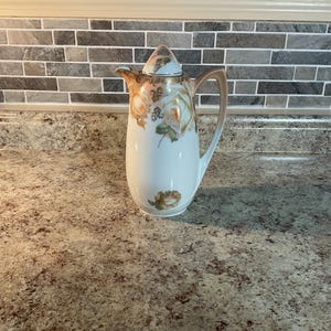 May include: White porcelain coffee pot with a floral design featuring white roses and green leaves. The pot has a lid with a gold trim.