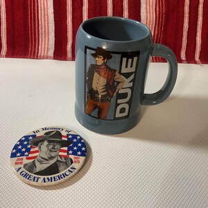 Puede incluir: Una taza azul con asa que presenta un gráfico de vaquero y la palabra "DUKE". Una posavasos con una imagen de John Wayne y el texto "In Memory of A Great American" está al lado de la taza.