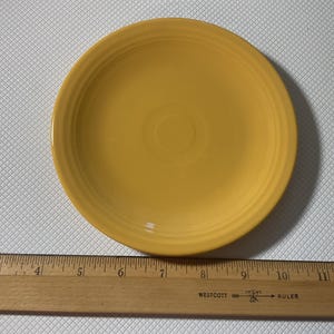 Può includere: Un piatto rotondo in ceramica gialla con anelli concentrici vicino al bordo. Il piatto ha un diametro di circa 23 cm. Un righello di legno è in primo piano.