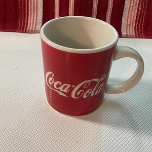 Puede incluir: Una taza roja de Coca-Cola con interior y asa blancos. El logotipo de Coca-Cola está en escritura blanca en la parte delantera. La taza está sobre una superficie blanca, con un fondo a rayas rojas y blancas. La taza mide aproximadamente 10 cm de alto.