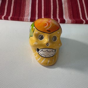 May include: A yellow ceramic skull figurine featuring a bright orange crown, green accents on the side, and white painted details around the eye sockets and jaw. The skull has hollow eye sockets, a nasal cavity, and a wide, grinning mouth with teeth.