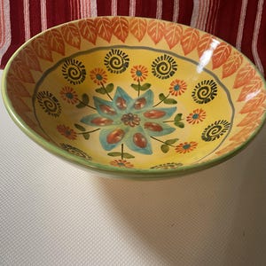 May include: A decorative ceramic bowl with a yellow interior, featuring a floral design in shades of blue, orange, and green. The rim is green, and the bowl has a leaf pattern around the edge. This bowl is suitable for serving food.