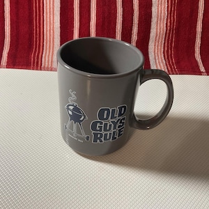 Może przedstawiać: Szary ceramiczny kubek z czarno-białą grafiką grilla i napisem "OLD GUYS RULE". Kubek ma zakrzywiony uchwyt i skośną krawędź. Kubek stoi na białej powierzchni z czerwono-białym paskowanym tłem.