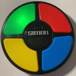 May include: A black electronic Simon game with four colored quadrants: green, red, yellow, and blue. The green quadrant is lit. The word "SIMON" is printed in silver on the center of the game.