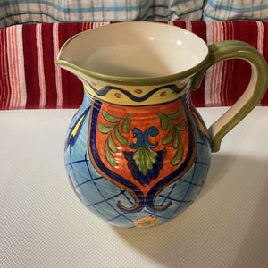 May include: A ceramic pitcher with a wide opening and a curved handle. The pitcher is painted with a blue, orange, and green floral design. The handle is green, and the interior is white.