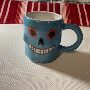 May include: A blue ceramic mug featuring a skull design. The skull has orange and pink flower accents around the eyes, a black nose, and a smiling mouth. The mug has a large handle and a white interior.