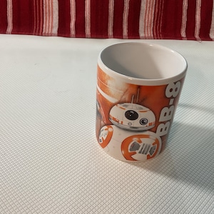May include: A white ceramic mug featuring the Star Wars character BB-8. The mug has an orange and white design with the BB-8 character prominently displayed. The mug is cylindrical and has a white interior.