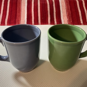Puede incluir: Dos tazas de cerámica. Una es de color azul grisáceo apagado, la otra es verde apagado. Ambas tienen un diseño curvo y un asa redondeada. Las tazas están sobre una superficie blanca con un fondo a rayas rojas y blancas.