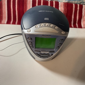 Peut inclure: Un radio-réveil lecteur CD Sony Dream Machine bleu foncé avec une bordure argentée. L'horloge a un écran vert et des boutons pour la fonction snooze. Les textes "DREAM MACHINE" et "SONY" sont visibles.