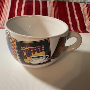 May include: A large, white ceramic coffee mug with a rounded handle. The mug features a colorful design with the words "Drink Coffee" and an illustration of a coffee cup and saucer. The background includes red, yellow, and green accents.