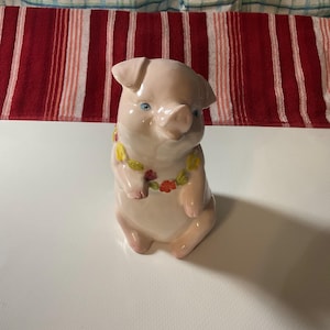 May include: A ceramic pig figurine in a seated position, with a light pink body and blue eyes. The pig wears a floral lei with yellow and red flowers. The background features a red and white striped fabric.