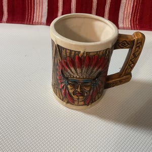 Pode incluir: Caneca de cerâmica vintage com um design de chefe nativo americano. A caneca apresenta uma base bege, uma alça marrom e uma imagem detalhada de um chefe com um cocar de penas em vermelho, azul e marrom.