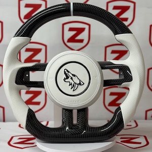 Puede incluir: Un volante de coche en blanco y negro con un diseño de fibra de carbono. El volante tiene empuñaduras de cuero blanco con costuras negras y un centro blanco con un logotipo de lobo. El fondo es un patrón de escudos rojos con la letra "Z".