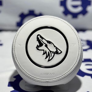 May include: White steering wheel featuring a black wolf emblem. The wheel has a stitched leather look, with the word "AIRBAG" printed at the bottom. The emblem is a detailed black illustration of a wolf's head, set within a black circle.
