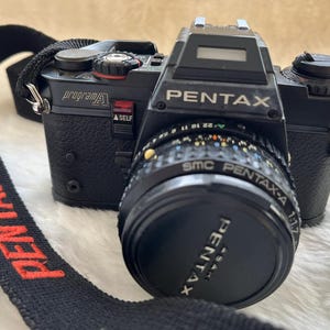 Könnte beinhalten: Schwarze Pentax Program A Kamera mit Objektivdeckel und schwarzem Gurt mit rotem "PENTAX"-Text. Die Kamera hat eine silberne Oberseite und ein Objektiv mit verschiedenen Farbkennzeichnungen. Das Objektiv lautet "SMC PENTAX-A 1:1.7".