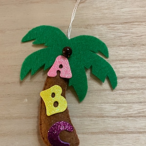 Pode incluir: Um enfeite de feltro em forma de palmeira com folhas verdes, tronco castanho e letras A, B e C com glitter. As letras são rosa, amarelas e roxas. Uma pequena conta castanha está no topo, e um cordão branco está preso para pendurar.