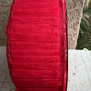 Op de afbeelding: Een rol helder rood cr&ecirc;pepapier, strak opgerold. Het papier heeft een textuur, gekreukte uitstraling, met zichtbare horizontale lijnen. De randen van de rol zijn zwart. Het papier wordt waarschijnlijk gebruikt voor knutselen of decoratie.