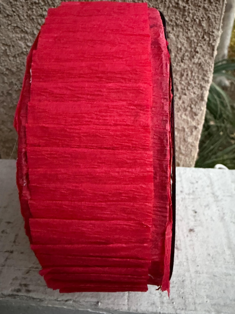 Op de afbeelding: Een rol helder rood cr&ecirc;pepapier, strak opgerold. Het papier heeft een textuur, gekreukte uitstraling, met zichtbare horizontale lijnen. De randen van de rol zijn zwart. Het papier wordt waarschijnlijk gebruikt voor knutselen of decoratie.