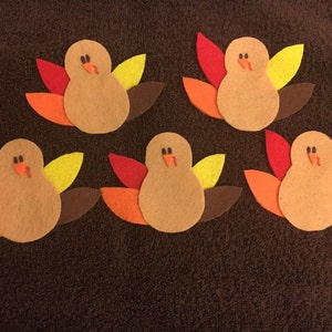 May include: Five felt turkey cutouts arranged in a group. Each turkey has a tan body, two black eyes, an orange beak, and a red wattle. Behind each body are four felt feathers in red, orange, yellow, and brown, fanned out in a semi-circle.
