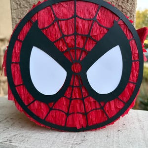 Op de afbeelding: Een ronde Spider-Man pi&ntilde;ata met een rode cr&ecirc;pepapier achtergrond en een zwart-wit maskerontwerp. De zwarte maskeromtrek heeft een webpatroon en grote witte oogvormen.