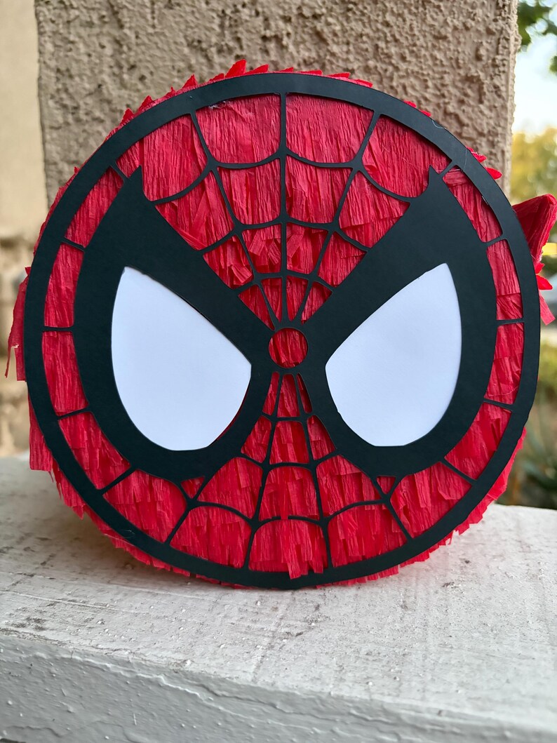 Op de afbeelding: Een ronde Spider-Man pi&ntilde;ata met een rode cr&ecirc;pepapier achtergrond en een zwart-wit maskerontwerp. De zwarte maskeromtrek heeft een webpatroon en grote witte oogvormen.