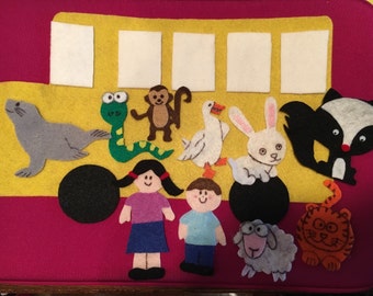 May include: A felt school bus with a yellow body and pink base, featuring white windows. Felt animal and child figures include a seal, snake, monkey, goose, rabbit, skunk, tiger, sheep, and two children. A black wheel is on the side.