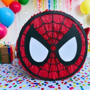 Op de afbeelding: Een ronde, rode Spider-Man pi&ntilde;ata met een zwart webdesign en witte ooguitsparingen. De pi&ntilde;ata is versierd met rood cr&ecirc;pepapier en is klaar voor een verjaardagsfeestje. Kleurrijke ballonnen en confetti zijn op de achtergrond.