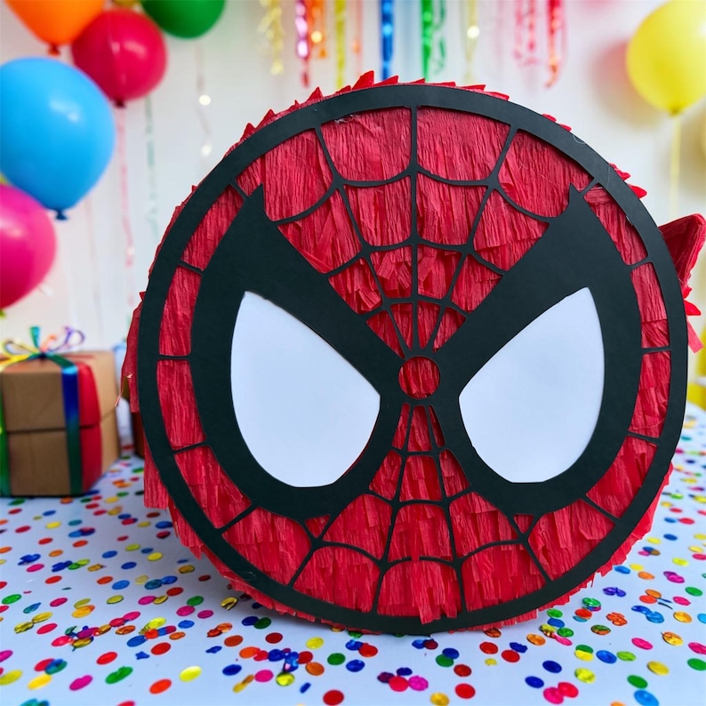 Op de afbeelding: Een ronde, rode Spider-Man pi&ntilde;ata met een zwart webdesign en witte ooguitsparingen. De pi&ntilde;ata is versierd met rood cr&ecirc;pepapier en is klaar voor een verjaardagsfeestje. Kleurrijke ballonnen en confetti zijn op de achtergrond.