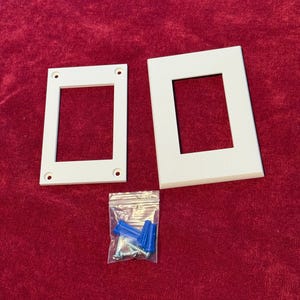 May include: Two white plastic wall plates with rectangular openings for electrical outlets. A small plastic bag containing four blue plastic anchors and four silver screws is included.