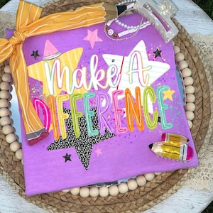 Può includere: Una t-shirt viola con il testo "Make A DIFFERENCE" in caratteri colorati e stilizzati. Il design include grafiche a stella gialle, bianche e nere con schizzi di vernice. Un nastro giallo con motivo a matita è legato sul lato.