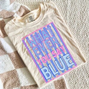 May include: Cream-colored t-shirt with the text "WHATEVER YOU SAY BLUE" in a blue and pink design. The shirt is folded and resting on a patterned blanket.